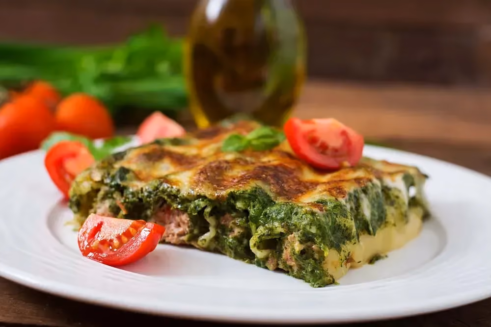Posé sur une assiette blanche, un pavé de cannelloni, épinards et boeuf mélangé est gratiné sur le dessus. Des tomates et de l'huile sont à côté.