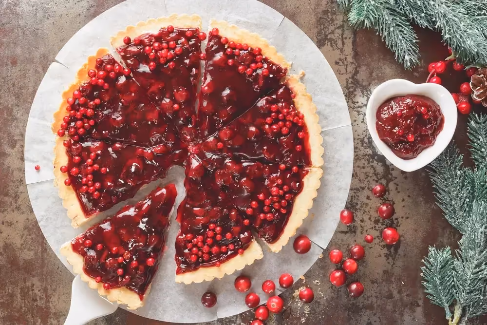 Sur une assiette blanche, le cheesecake aux canneberges est coupé en huit parts. Des branches d'épicéa et des petites baies rouges sont visibles.