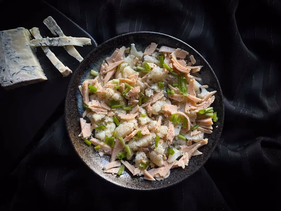 A small blue bowl holds cauliflower with gorgonzola and turkey breast. Some gorgonzola is placed next to the dish.