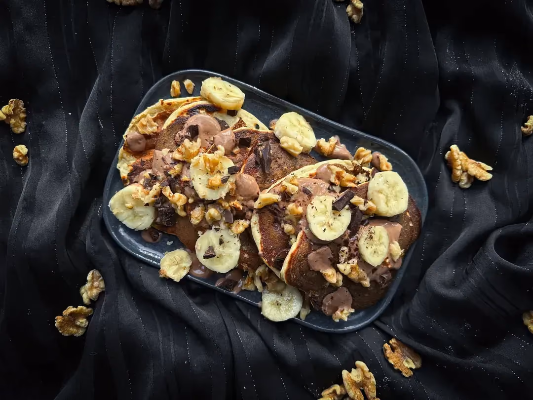 Un plat rectangulaire peu profond aux coins arrondis accueille quatre pancakes accompagnés de banane, chocolat noir et cerneaux de noix.