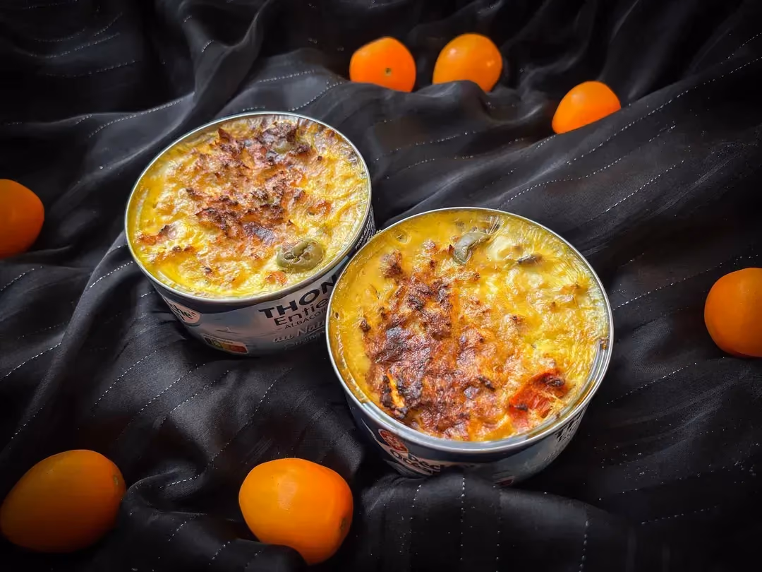 Deux boites de thon servent de réceptacle à la recette de clafoutis au thon, tomates, curry et olives vertes. Des tomates cerises sont à côté du plat.