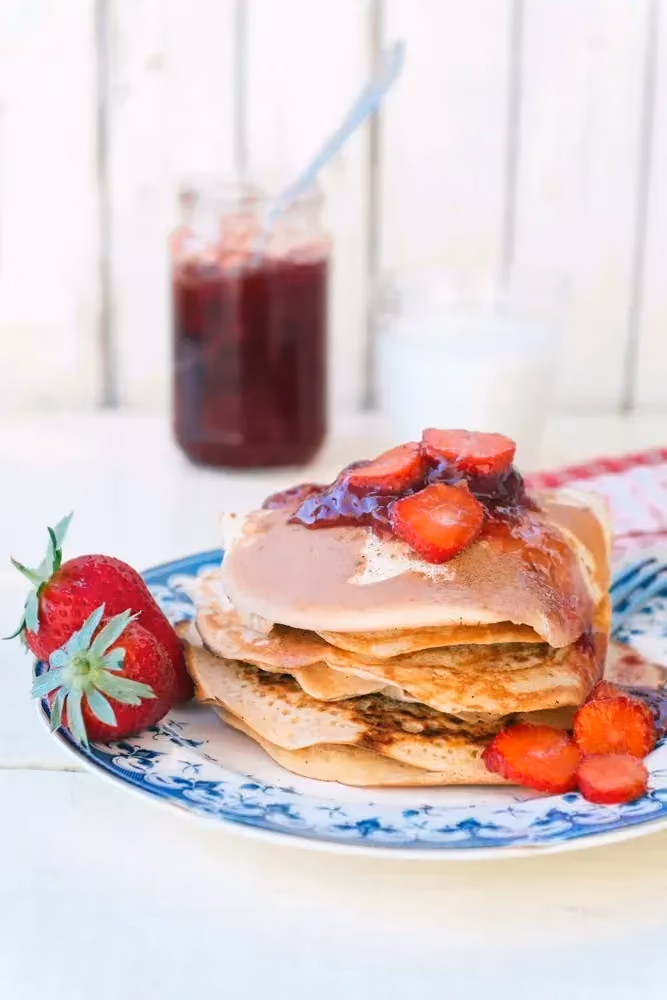 Un pot de confiture de fraises ouvert avec une cuillère se trouve à coté d'une assiette avec des pancakes et des fraises entières et en tranches.
