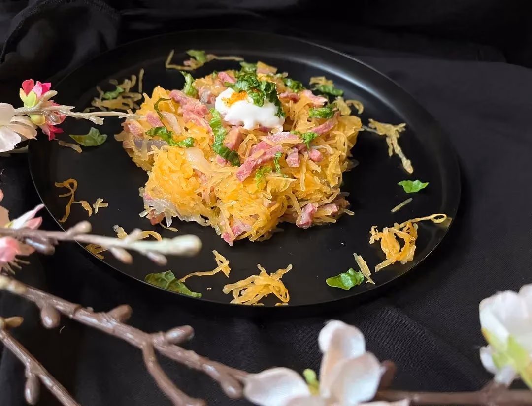 In the center of a black plate is spaghetti squash, cut into spaghetti-like strands, with bacon strips and crème fraîche on top.