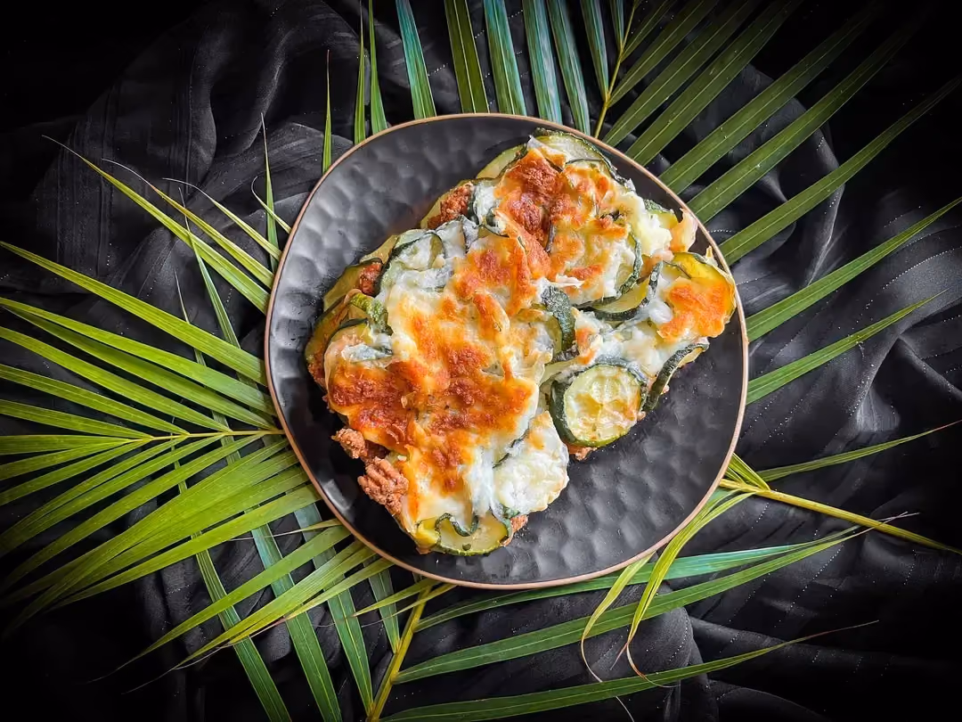 Sur du feuillage se trouve un petit saladier contenant les courgettes recouvertes de sauce Béchachel et de fromage râpé gratiné/