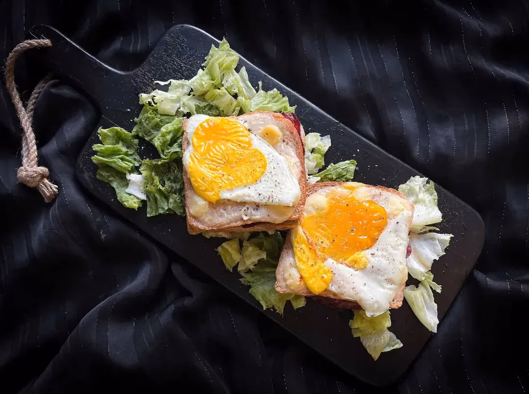 Two croque madames with viande des Grisons, béchamel sauce, and button mushrooms are placed on a ceramic board.