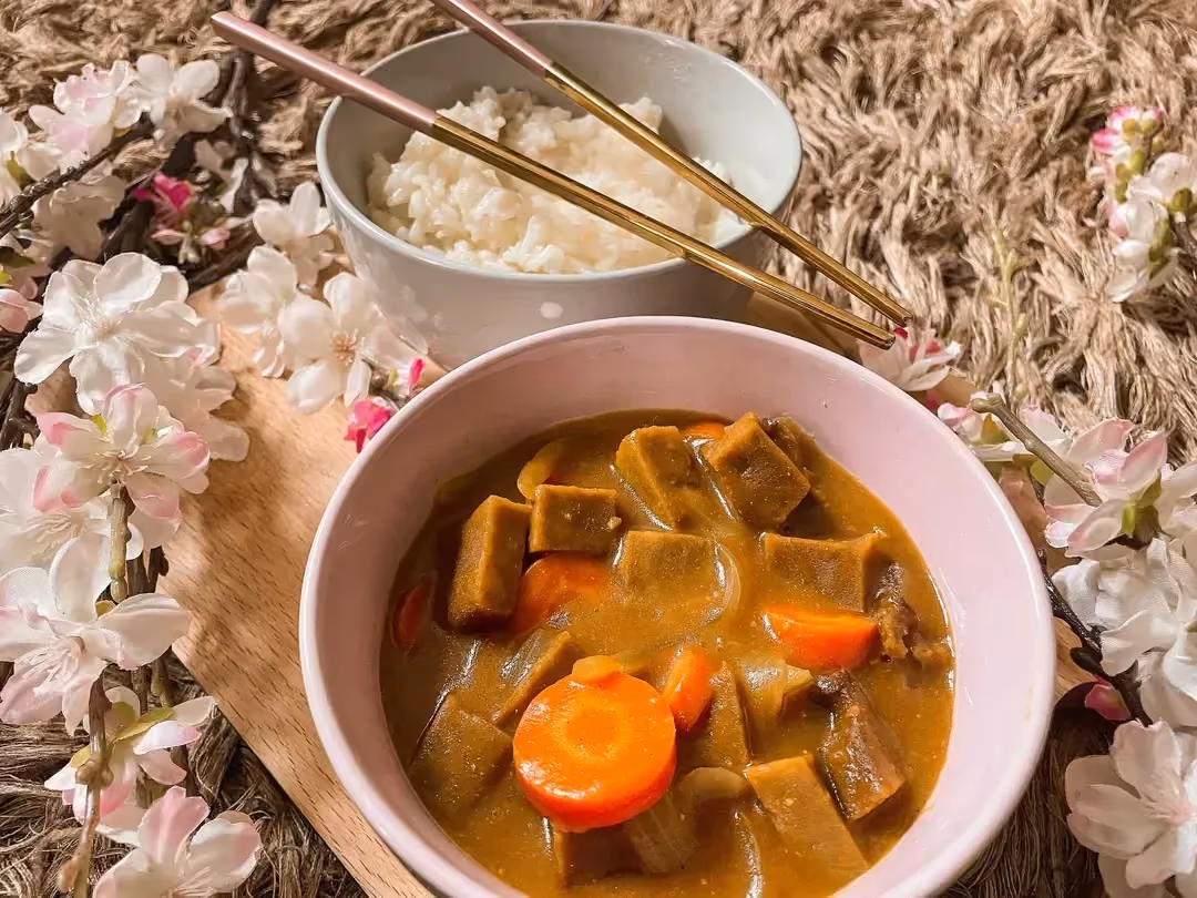 Le curry de seitan et carottes à la japonaise est accompagné d'un bol de riz à l'arrière plan, tout autour du curry il y a des fleurs de cerisier.
