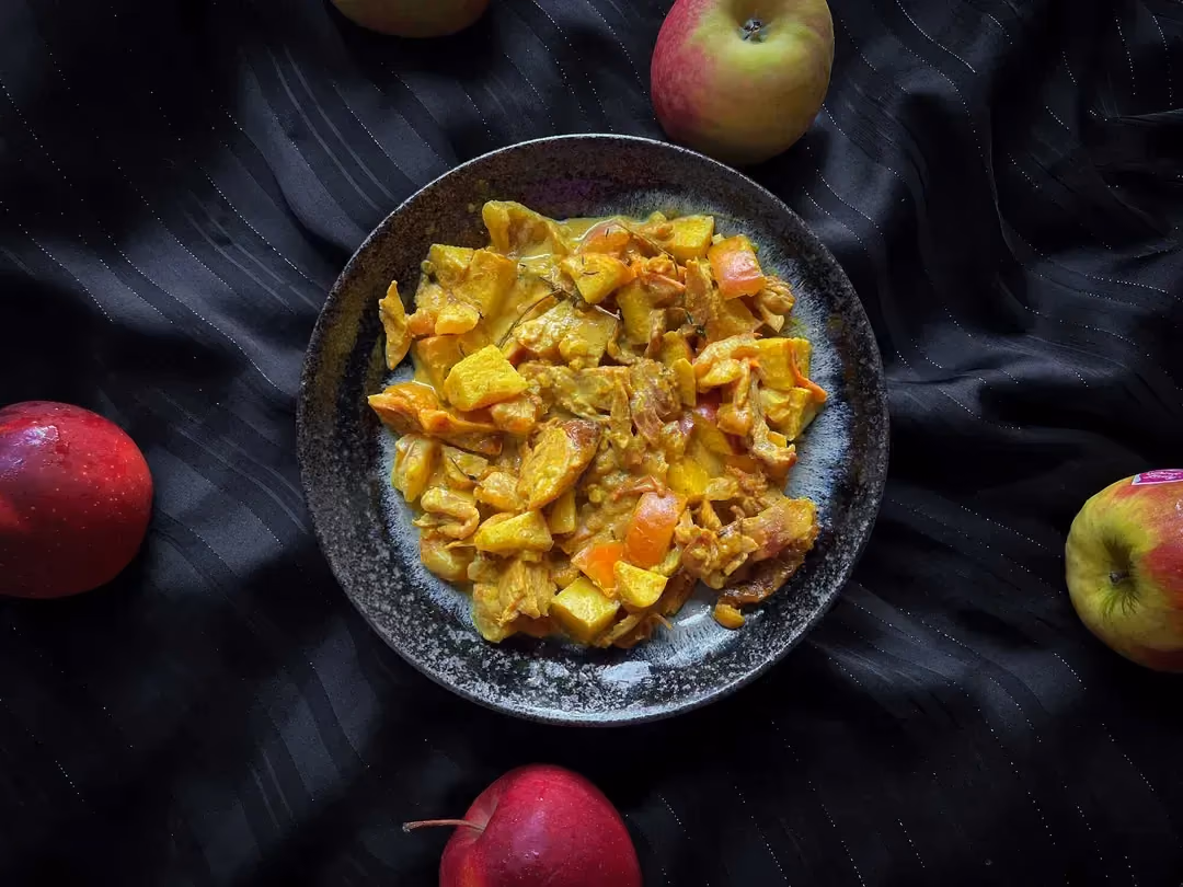 The red apple chicken curry recipe is presented in a small blue bowl. Four apples are arranged around the dish.