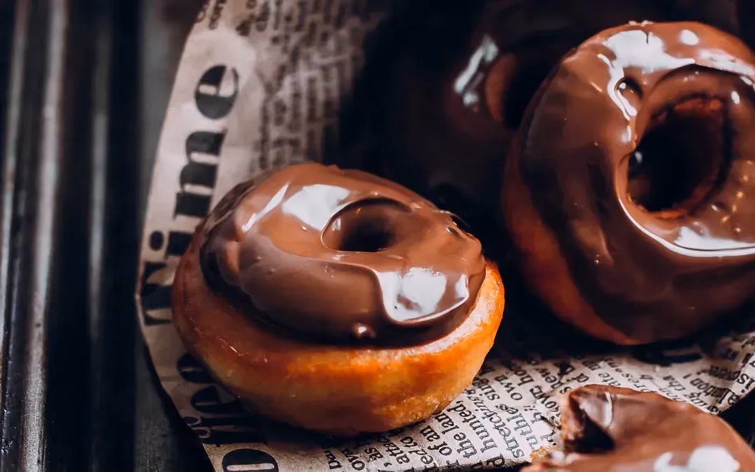Sur une feuille de journal sont présentés trois donuts recouvert du glaçage au chocolat. L'un des trois est déjà entamé.