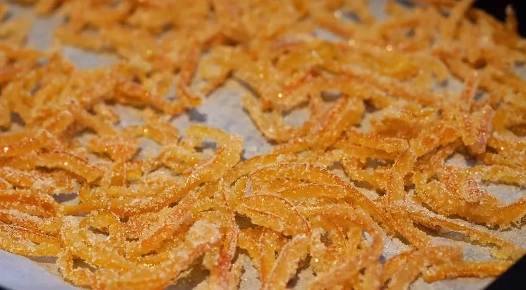 Une multitude de fines lamelles d'écorces d'oranges confites enrobées de sucralose sont déposées sur une plaque de cuisson.