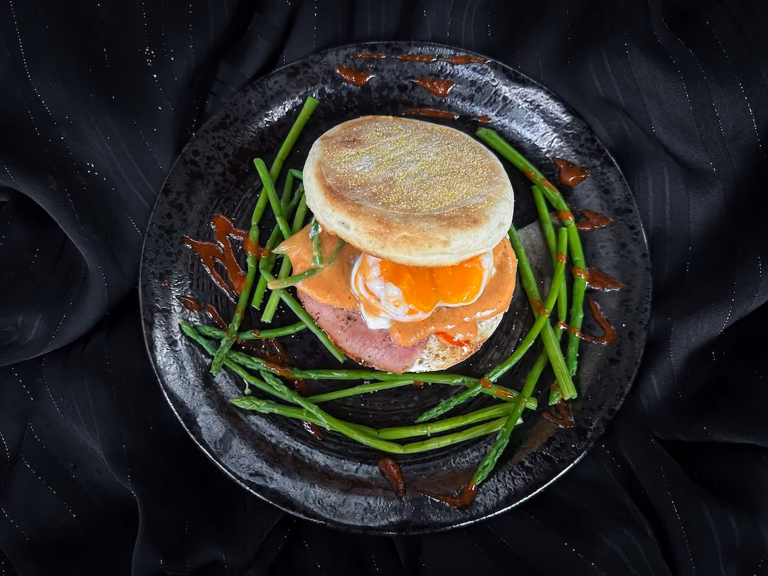 On a black plate is an English muffin topped with poached egg, bacon, a sriracha-mustard sauce, and Vache qui rit, served with asparagus.