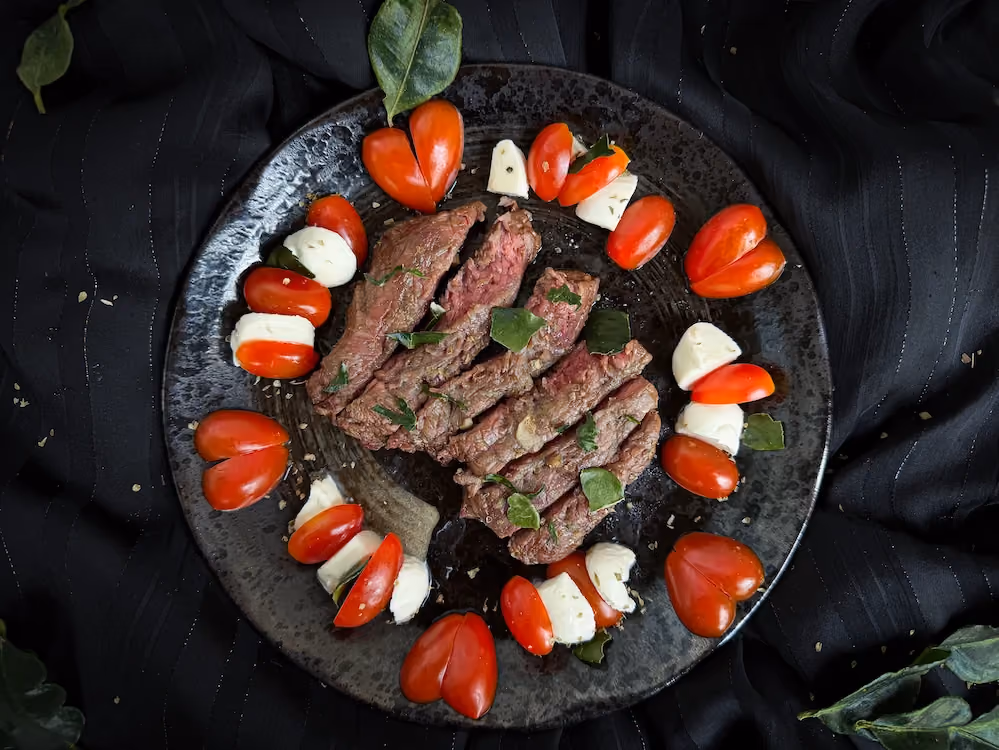 Sliced beef tenderloin is arranged in the center of the plate, garnished with basil, with cherry tomatoes and mozzarella around it.