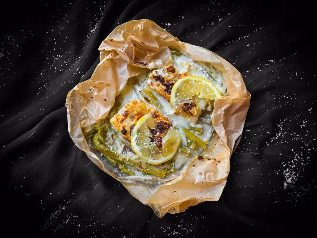 Two nicely seared cod fillets are served with small asparagus in a Parmesan cream sauce. Two small lemon slices are on top.