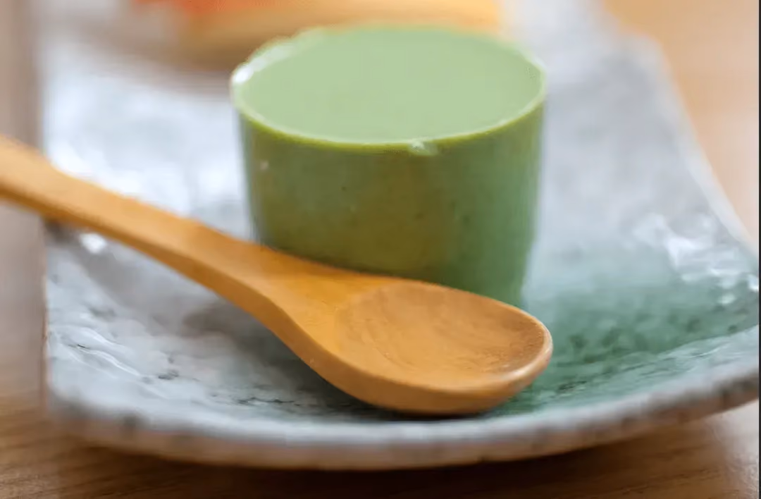 Le flan est dans une sorte de bol haut et étroit et il a pris la couleur de la pistache. Une grosse cuillère en bois se trouve à côté.