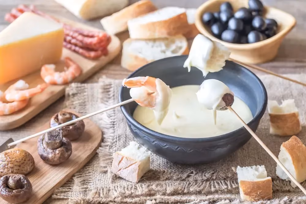 Placed on a table is a bowl of melted cheese. Shrimp, mushrooms, and slices of bread are also present.