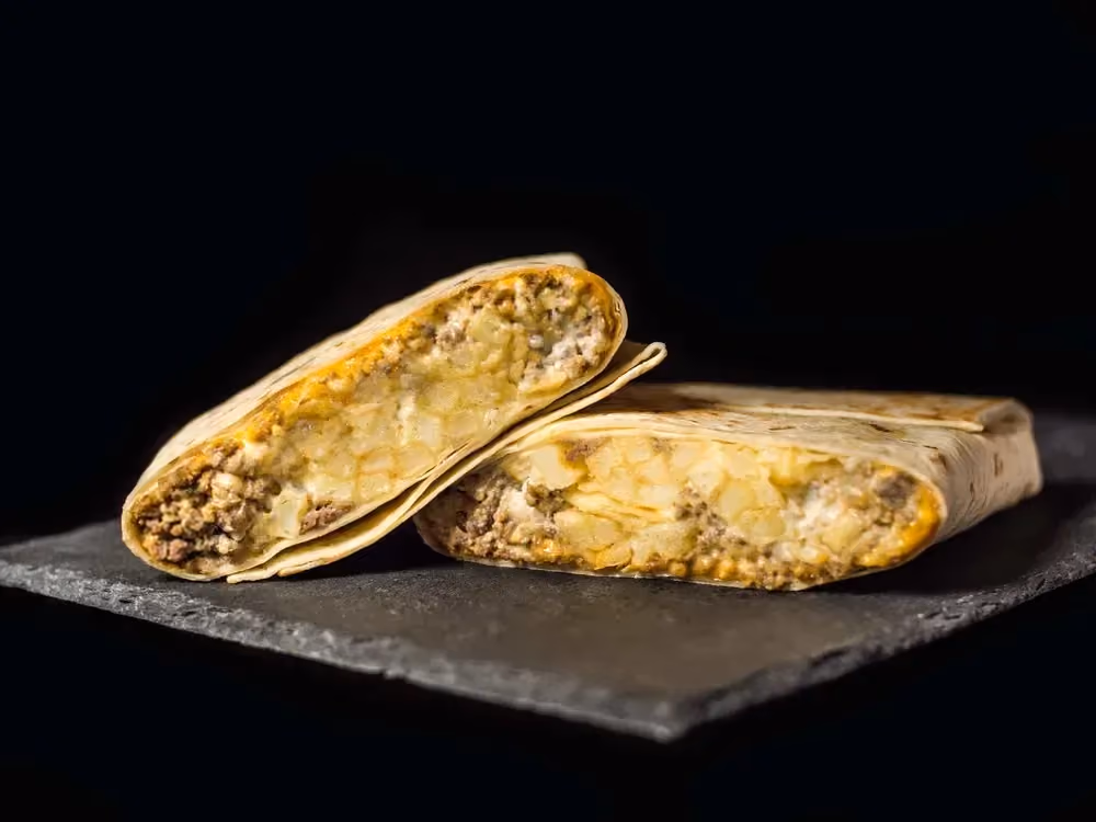 Deux gros French tacos à la viande de boeuf sont posé sur une plaque foncée. L'un des deux montre la généreuse garniture à base de viande.