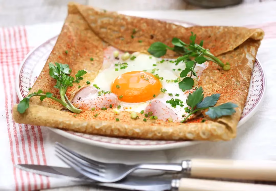 La galette bretonne, avec son oeuf sur le plat en son centre est présentée dans une assiette creuse en faïence blanche avec des motifs roses.