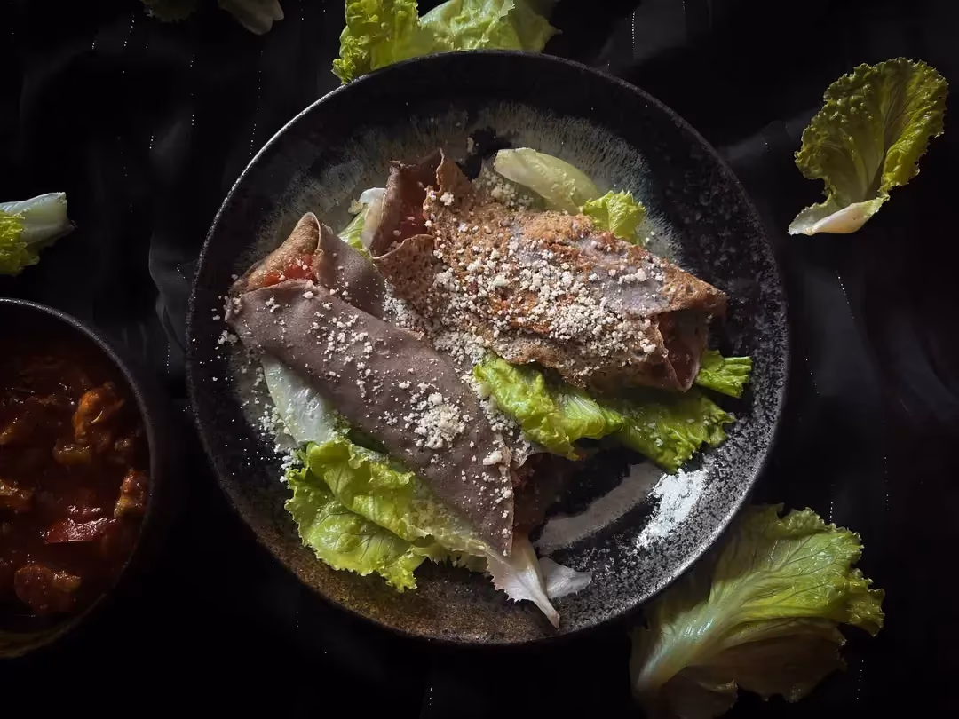 Two buckwheat galettes with Basque-style vegetables are served in a small bowl. A few lettuce leaves accompany the dish.