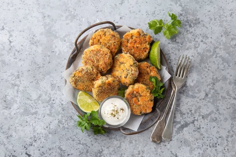 In a metal dish, there are eight pieces of salmon steak shaped like patties. Two lime wedges accompany the dish.