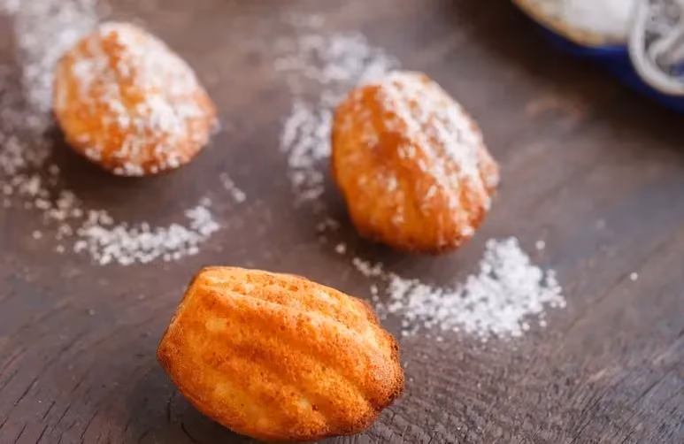 Sur un plan de travail de couleur sombre, se trouve trois jolies madeleines à la fleur d'oranger bien dorées et saupoudrées de poudre d'amande.