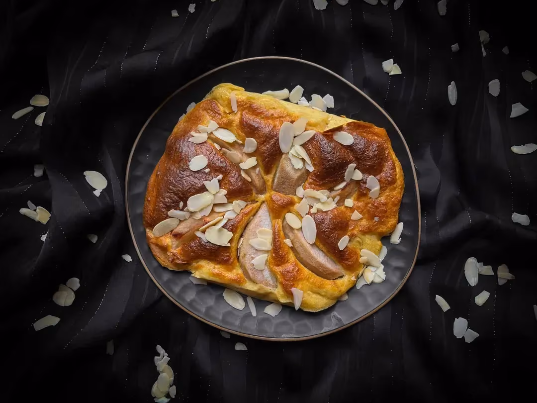 Le gâteau façon amandine aux poires est présenté dans un petit plat de couleur bleu. Sur le dessus et autour du plat se trouve des amandes effilées.