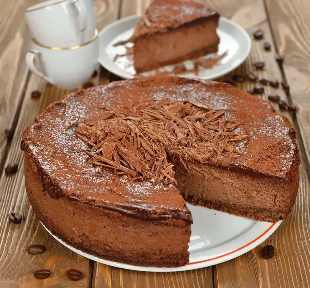 On a wooden table, there is a plate holding a chocolate cheesecake. A small slice has already been taken and placed on a small plate.