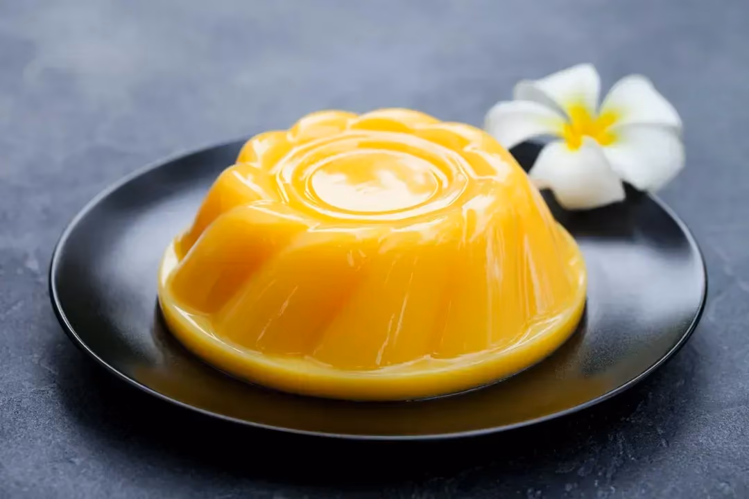 Cette gelée à la mangue ressemble à un flan. Elle est présentée sur une large assiette de couleur noire. Une fleur de tiaré complète l'ensemble.