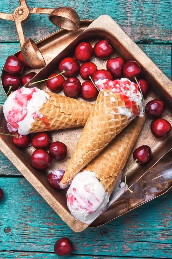 Sur une table en bois rustique, dans un plat de forme carré se trouve trois cornets de glace à la cerise accompagnés par des cerises entières.
