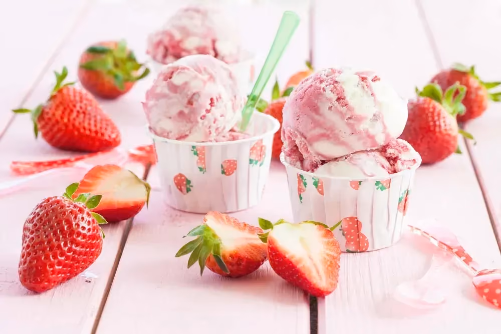 Deux petits gobelets contiennent de la glace à la fraise. A côté on voit des fraises entières et d'autres coupées en deux.