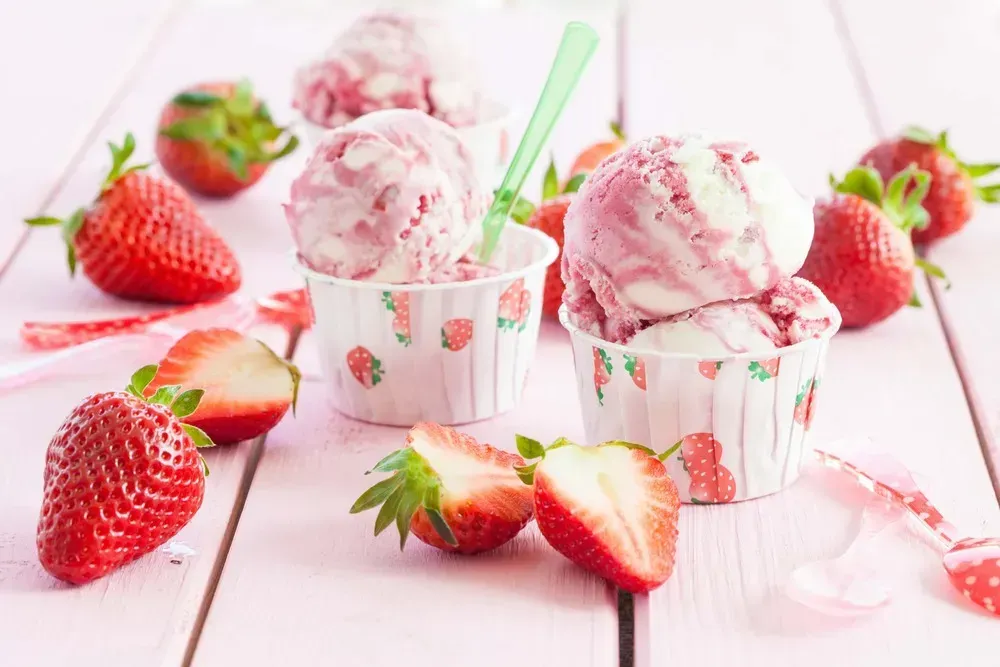 Two small cups contain strawberry ice cream. Next to them, you can see whole strawberries and others cut in half.