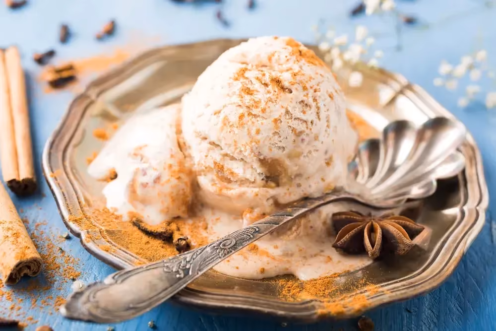 Une assiette en faïence de couleur beige contient une grosse boule de glace. Une grosse cuillère en argent en forme de feuille est à côté de la glace.