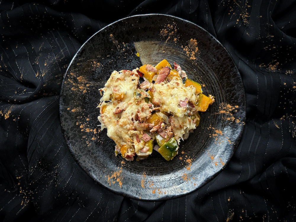 This squash gratin is presented in a white earthenware charlotte mold. On top is grated cheese and a silver pie server.