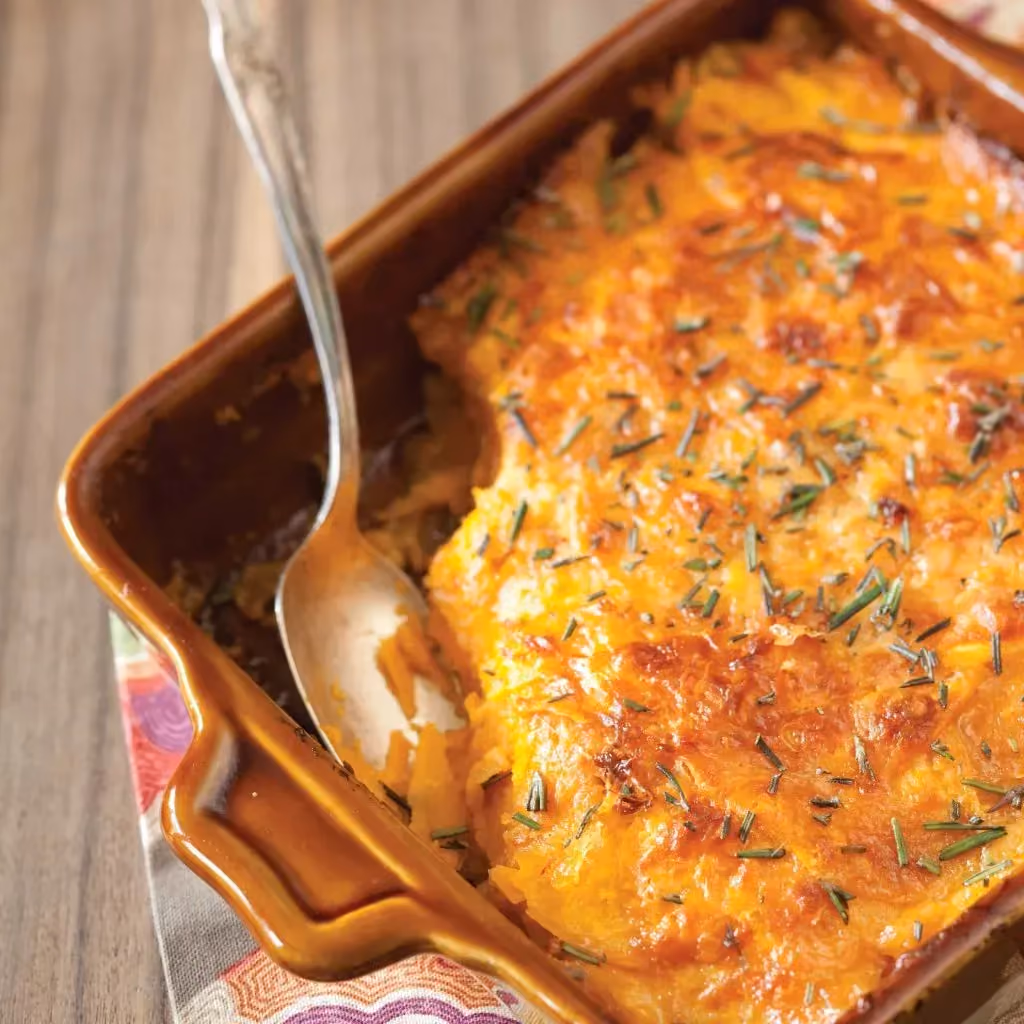 Set on a wooden table, a brown baking dish holds this haddock and sweet potato gratin. A generous portion has been served out of the gratin.