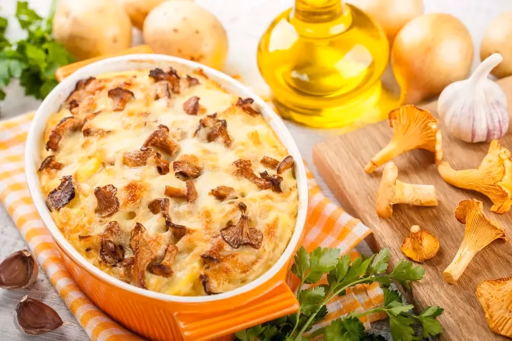 An orange plate holds this chicken, potato, and chanterelle gratin. Next to it are garlic, onion, and chanterelles.