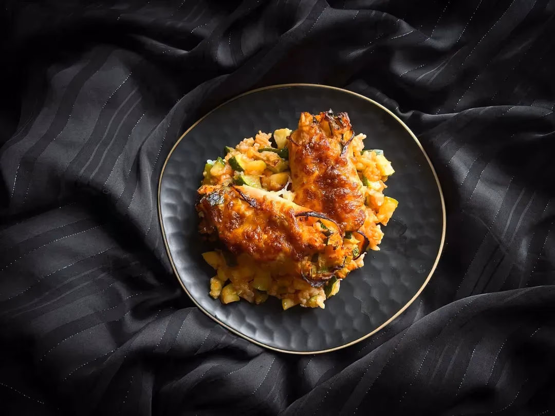 Two slices of baked ham are rolled around rice, zucchini, and tomato sauce.