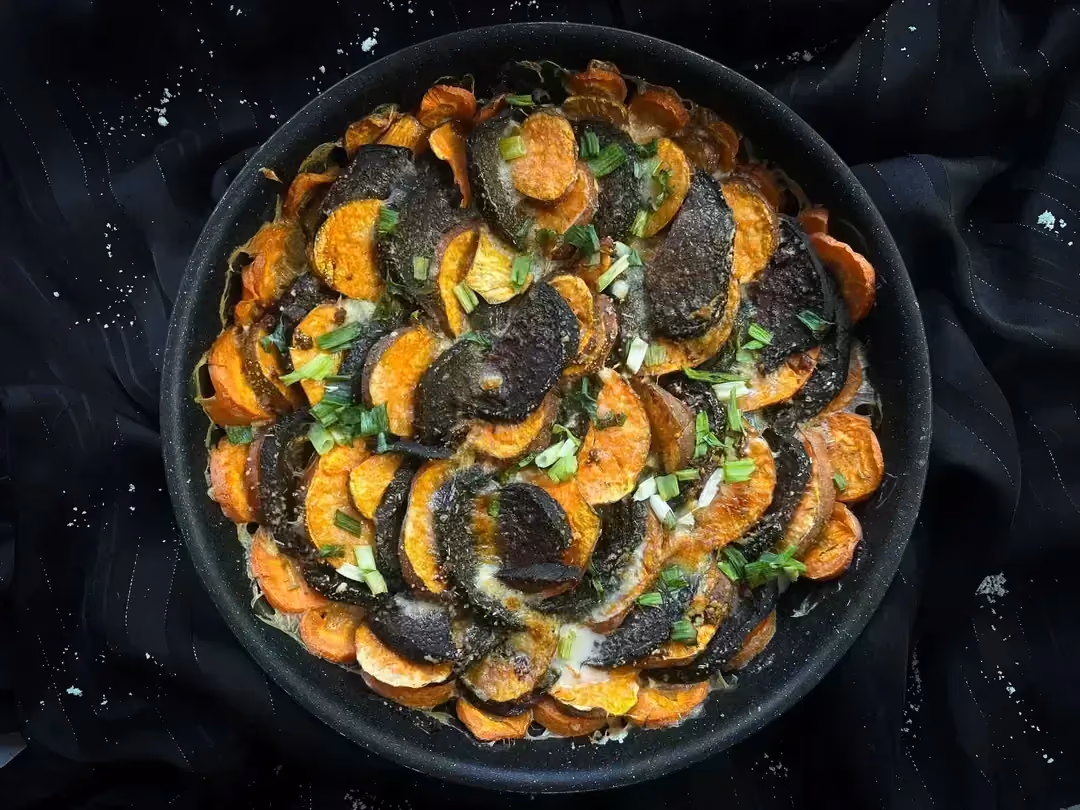 In the baking dish are the recipe ingredients, such as sweet potatoes, beets, and carrots with Parmesan.