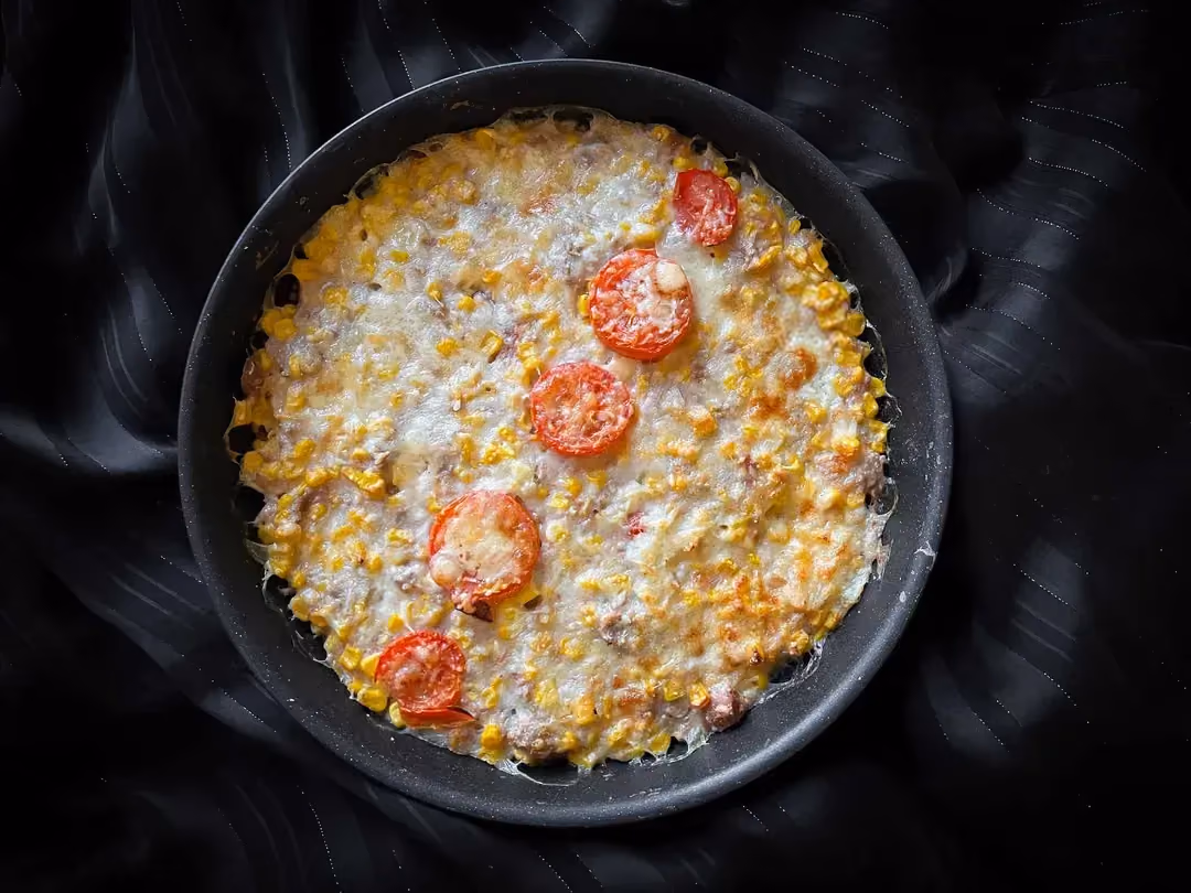 The tuna gratin with corn is presented in a blue bowl. 5 slices of grilled tomato are lined up on top of the gratin.
