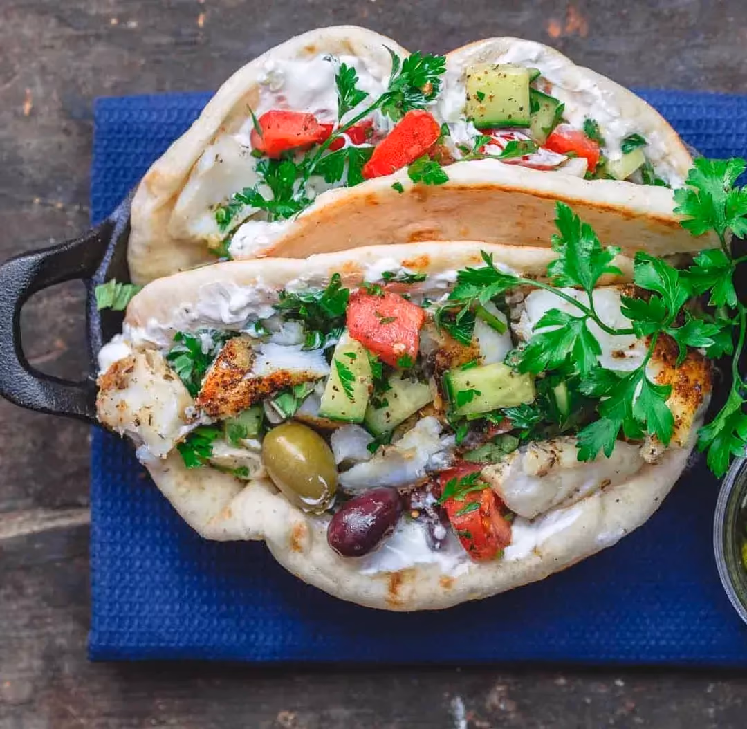 On slices of pita bread, there is grilled cod, and you can see tomatoes, cucumbers cut into small cubes, and olives.