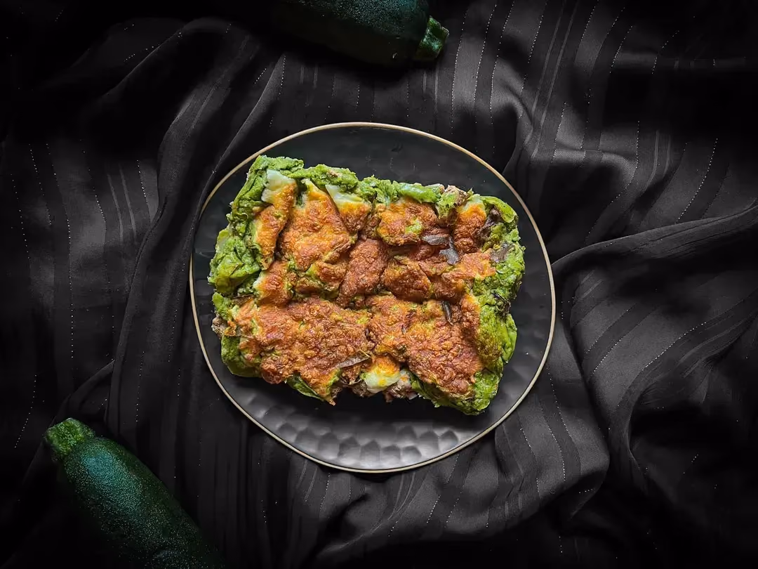 In a small blue textured bowl is shepherd's pie with green vegetables. A zucchini is placed next to the dish.