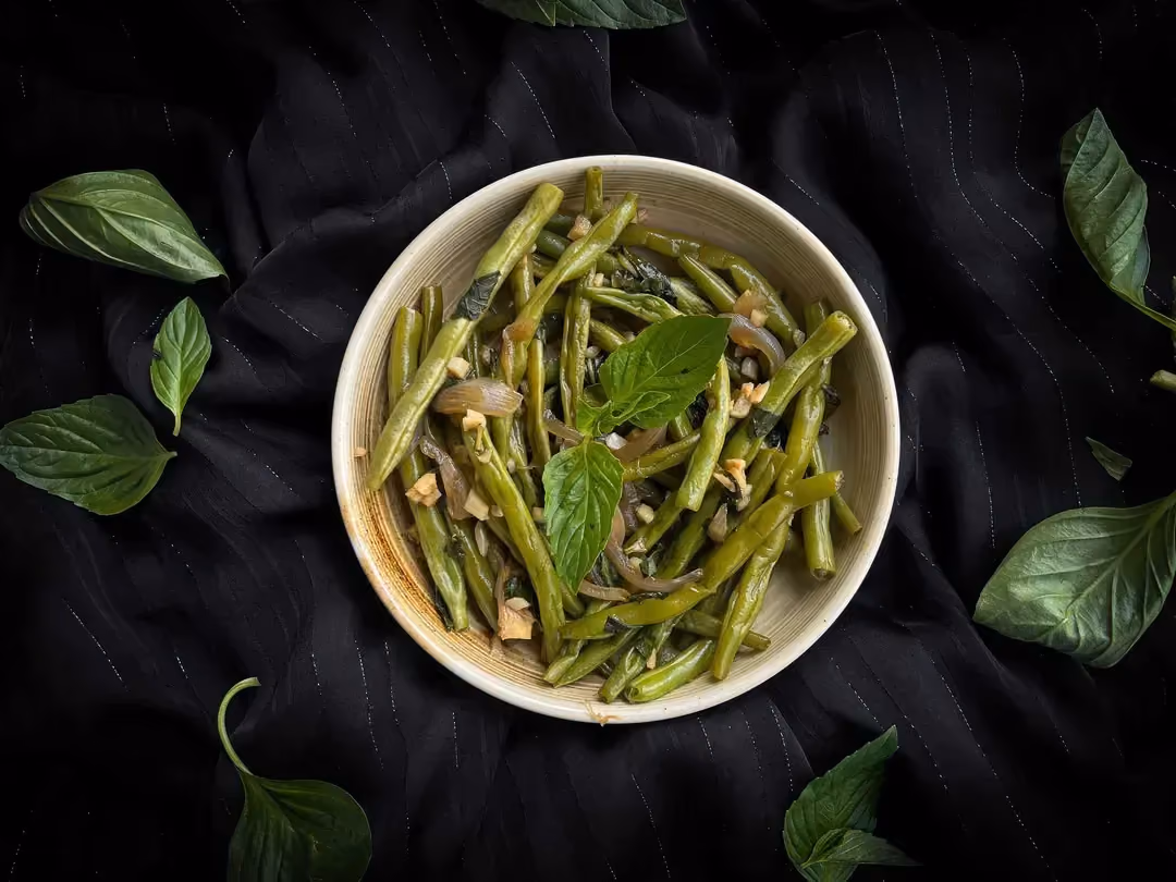 Dans un plat en faïence de couleur blanc, les haricots verts accompagnés d'ail et persil sont prêts à être consommés. Deux fourchettes sont visibles.