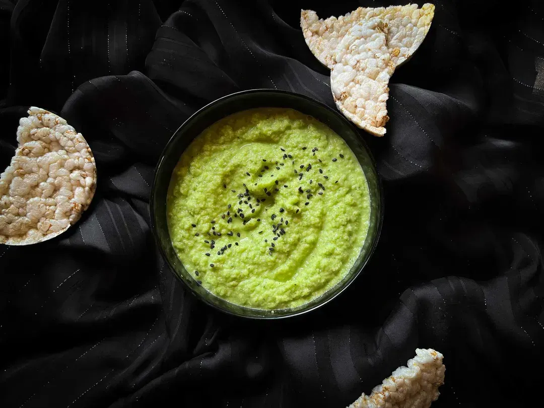Zucchini hummus sprinkled with sesame seeds is served in a small blue bowl. The green sauce looks beautiful.