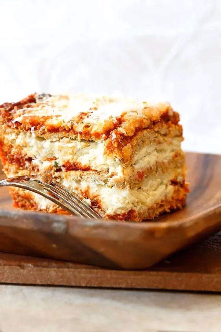 Une grosse part de lasagnes de chou-fleur est posée sur une assiette en bois. Une fourchette se trouve à côté en attente de dégustation.