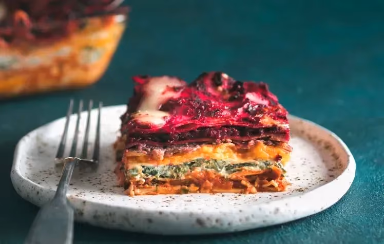 Dans une assiette de couleur blanche se trouve une grosse part de lasagnes multicolores tout juste sortie d'un plat à gratin.