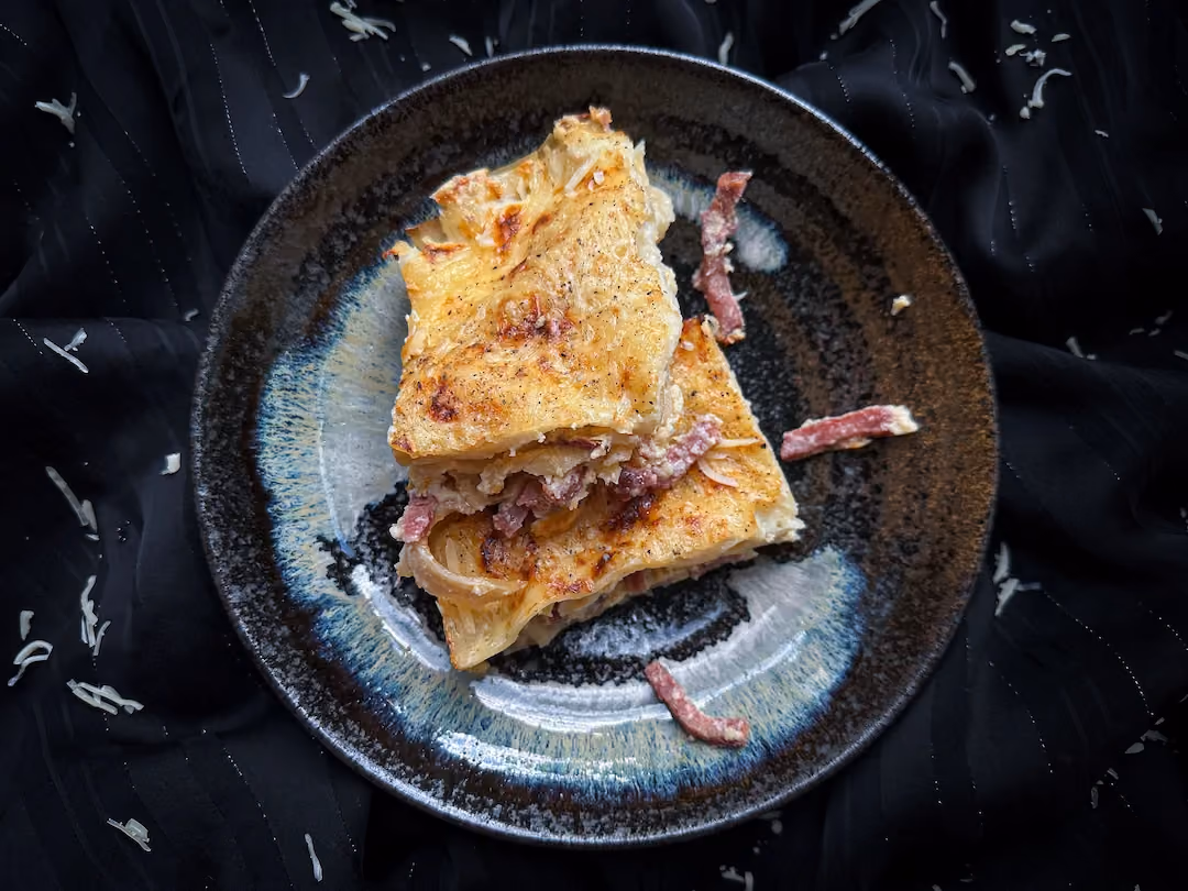 On a black background, there is a black plate holding carbonara-style lasagna.
