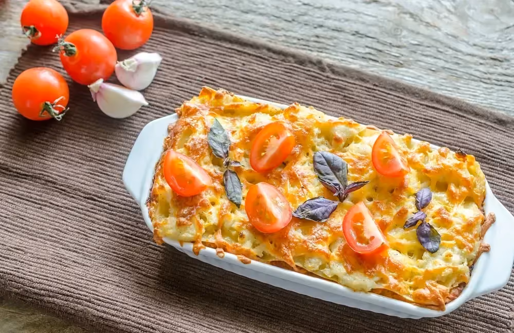 Un plat à gratin blanc rectangulaire accueille les lasagnes provençales. A côté se trouve quatre tomates cerises et deux grosses gousses d'ail.