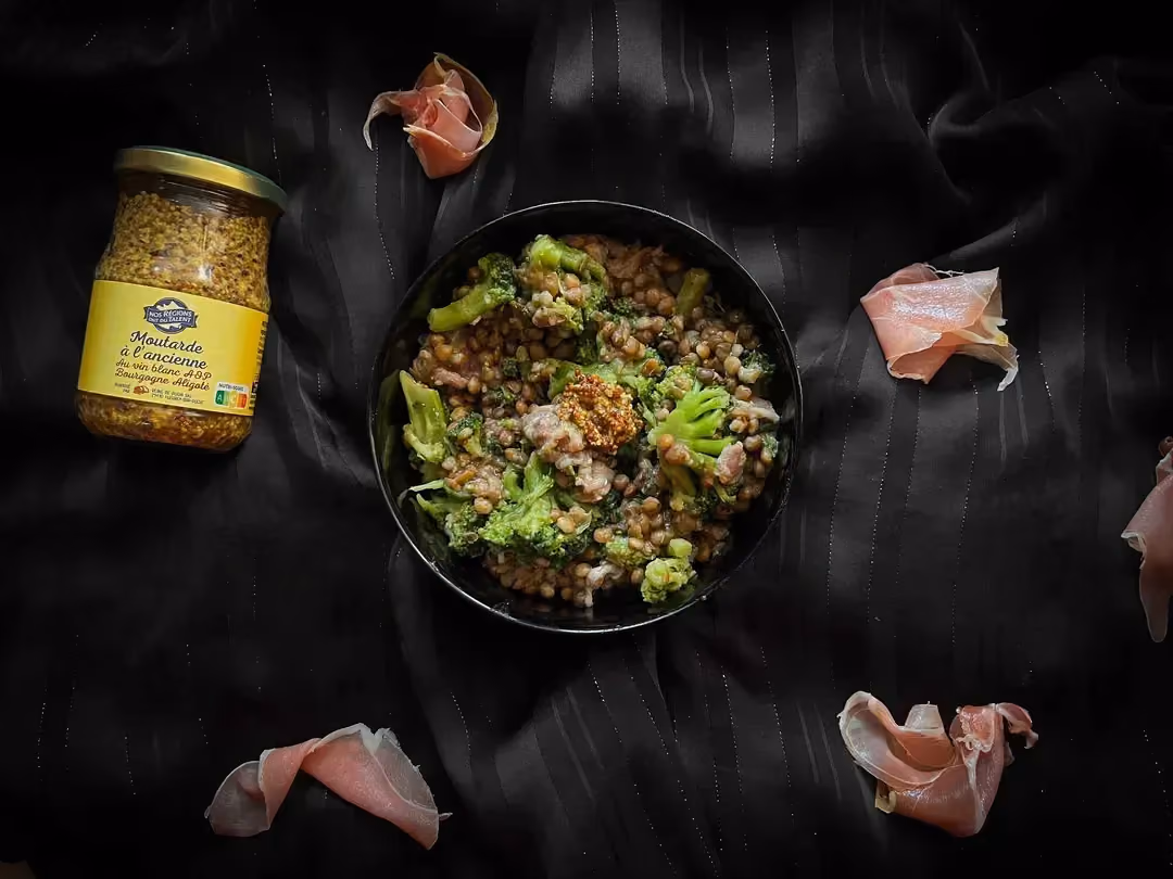 A small bowl holds the lentils, broccoli, Parma ham, pickle, and whole-grain mustard recipe. Next to it is a jar of mustard.