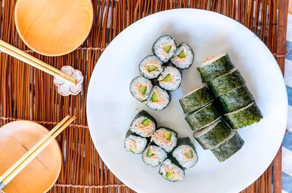 Dans une assiette creuse de couleur blanche se trouve une vingtaine de makis de thon. Deux petits bols vides et des baguettes sont à côté.s