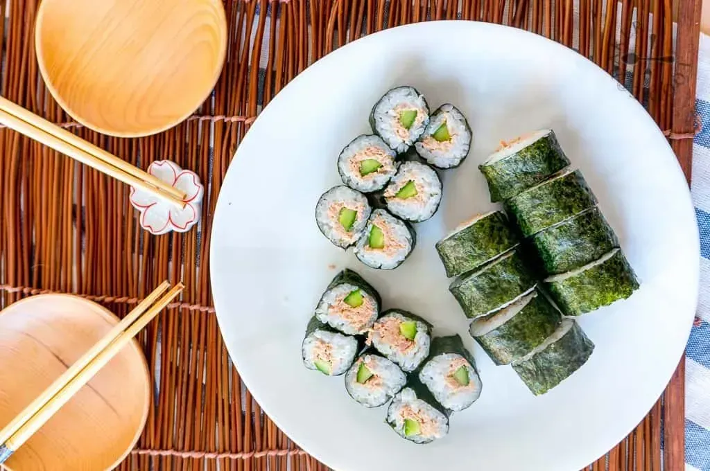 In a deep white plate, there are about twenty tuna maki rolls. Two small empty bowls and chopsticks are next to it.