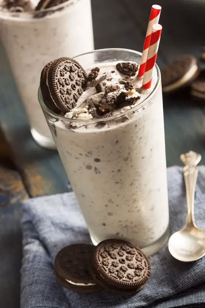 Deux grands verres remplis de milkshake protéiné sont prêt à être consommé. Deux pailles et deux biscuits Oréo trempent dans chacun des milkshakes.