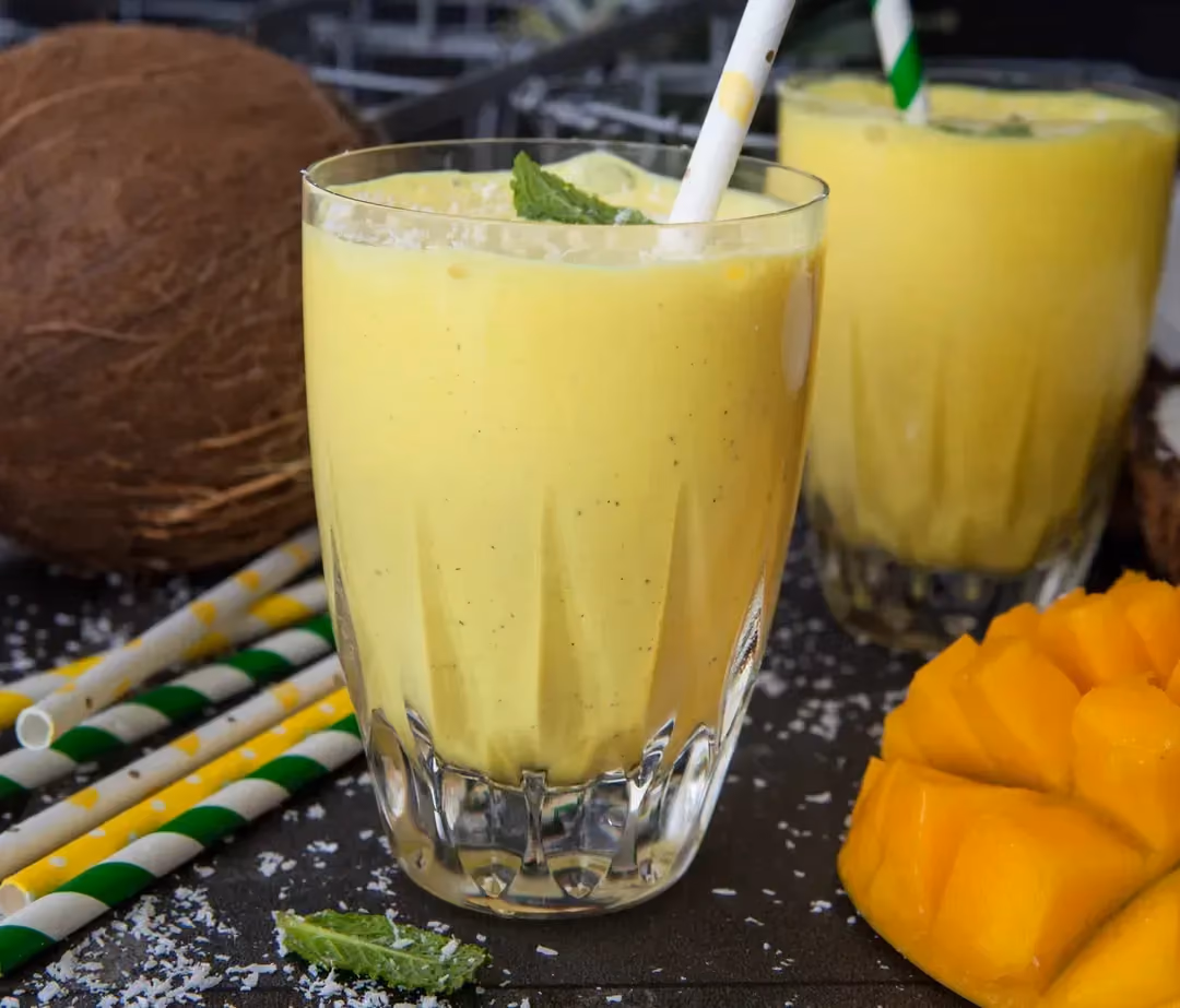 Le milkshake protéiné tropical est présenté dans deux grands verres avec des pailles. Une grosse mangue sans sa peau à la chair jaune est à côté.