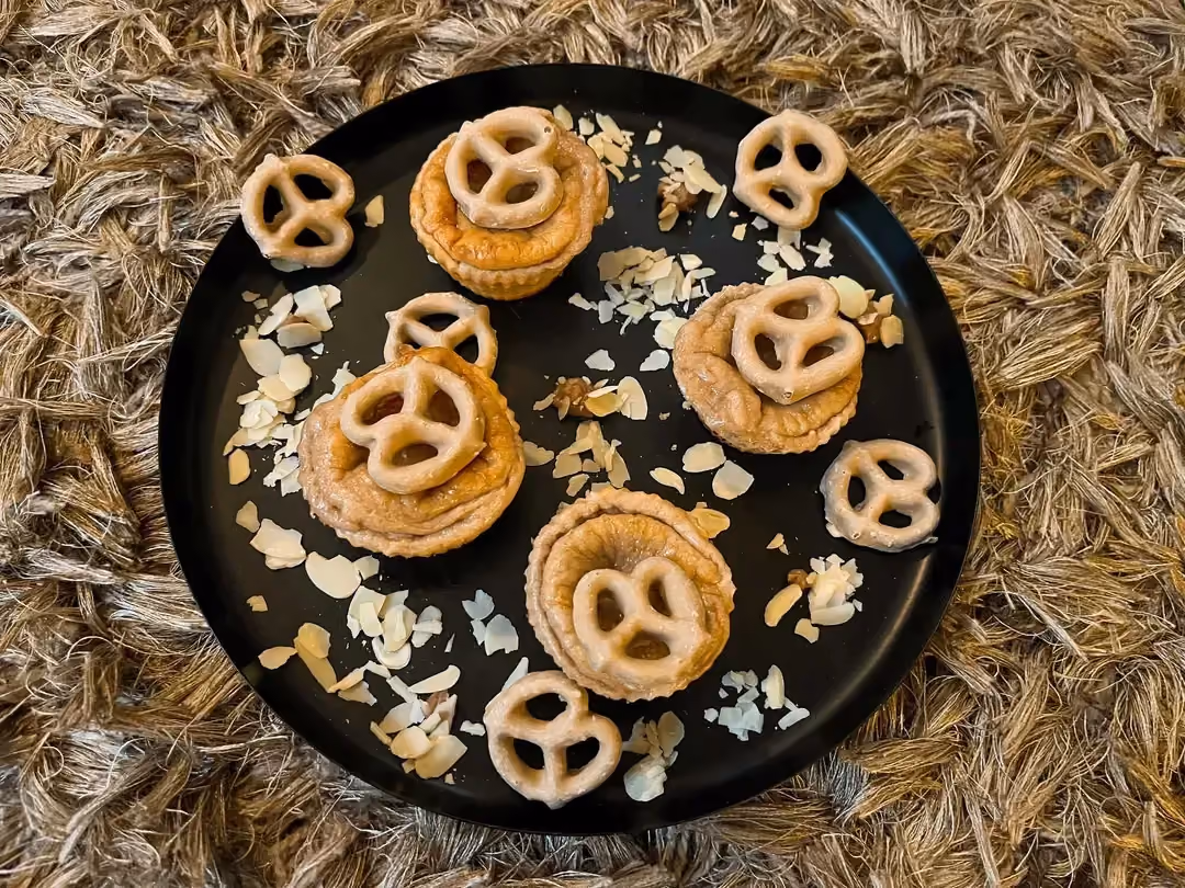 On a woven raffia mat, there is a pan holding four beautiful peanut muffins. Crushed peanuts are scattered around the muffins.